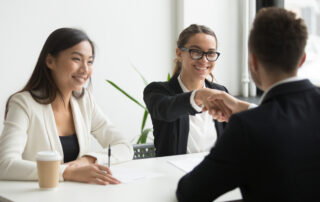 Male lawyer shaking the hand of female lawyer across the table. Another member of counsel is also sitting at the table smiling.