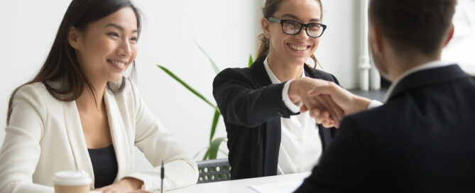 Male lawyer shaking the hand of female lawyer across the table. Another member of counsel is also sitting at the table smiling.